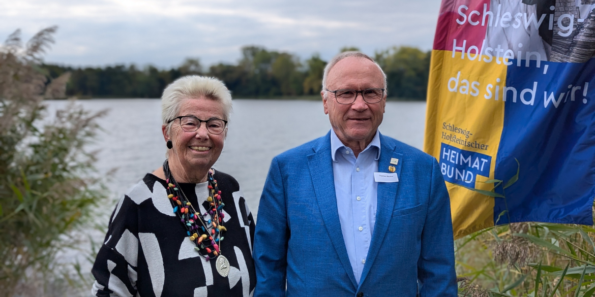 Zwei Personen vor See und Beachflag mit Aufschrift "Schleswig-Holstein, das sind wir!"