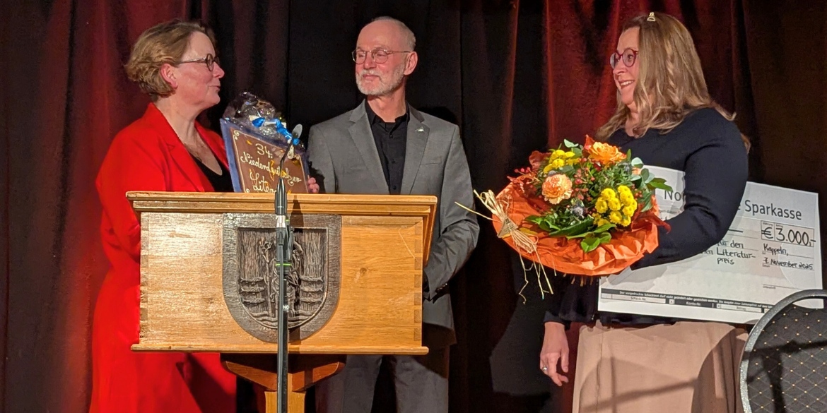 Drei Personen auf Bühne mit Blumen, Scheck und Schokolade