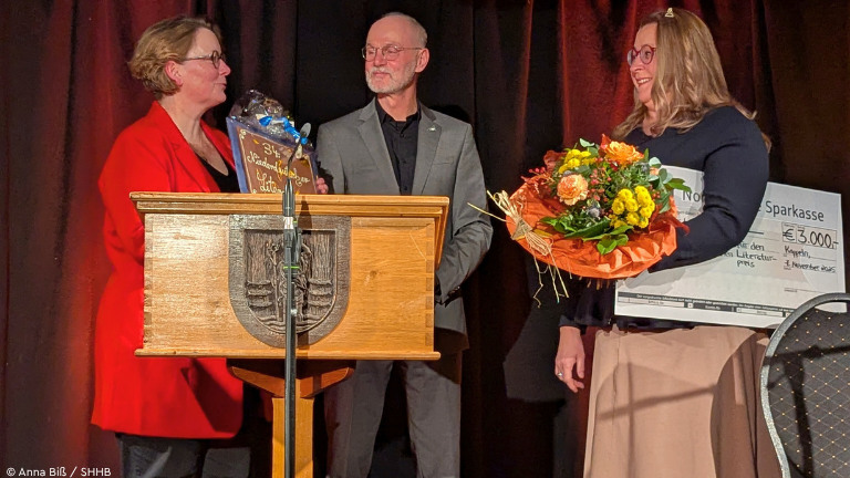 Drei Personen auf Bühne mit Blumen, Scheck und Schokolade