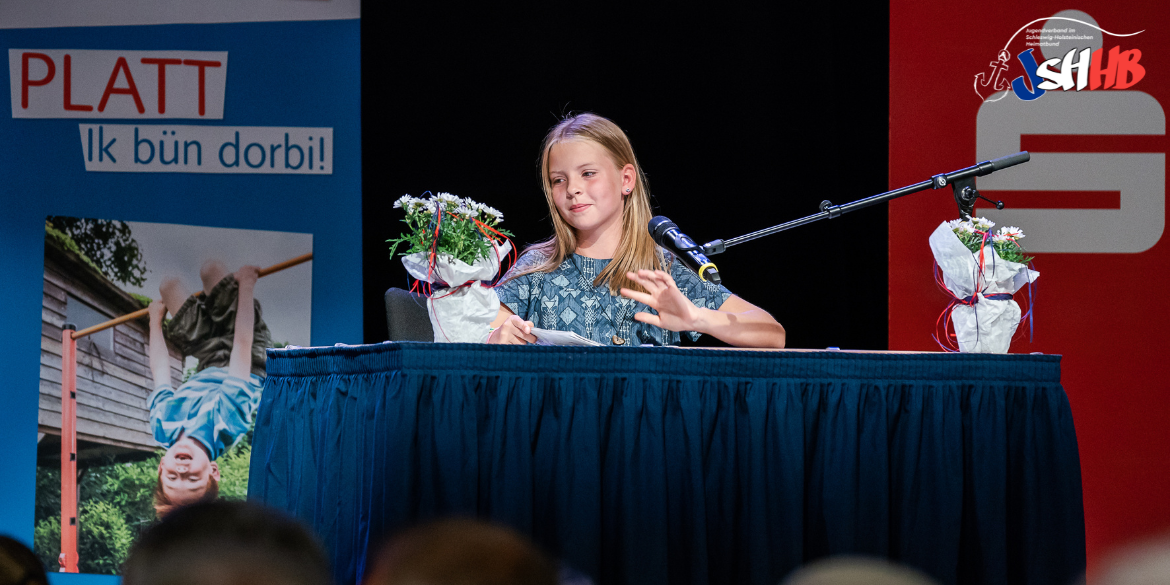 Ein Mädchen sitzt am Tisch auf einer Bühne und trägt etwas vor © SHHB