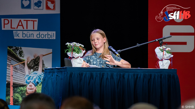 Ein Mädchen sitz am Tisch auf einer Bühne und trägt etwas vor